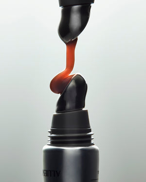 A close-up studio shot of a black cosmetic tube with a twisted applicator dispensing a glossy orange-red gel or cream upward against a light gray background.